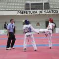 Duas atletas de taekwondo em luta sobre tatame. Ao lado esquerdo está o árbitro. #Pracegover