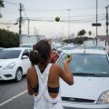 menina de costas para a foto faz malabares com limões na avenida diante de carros. #paratodosverem