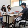 homem está em cadeira de rodas com curativos nos dois pés. Ele está diante de mesa. atrás da mesa estão sentadas duas mulheres. #paratodosverem
