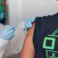 Imagem em close mostra mãos usando luvas vacinando um homem que está de costas. #Paratodosverem