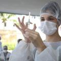 Mulher vestindo avental, usando touca e máscara manipula seringa de vacinação. #Paratodosverem