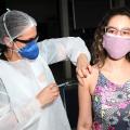 mulher usando avental, máscara e gorro vacina outra mulher. #paratodosverem