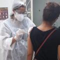 Mulher paramentada com avental, luva, gorro e máscaras vacina outra mulher. #Paratodosverem
