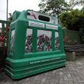 grande caixa de material plástico com abertura no alto para depósito de vidros. Ela está no chão de um parque, sobre solo pavimentado. #paratodosverem