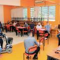 Sala de aula com cadeiras dispostas em círculo. Pessoas estão sentadas distantes umas das outras. #paratodosverem