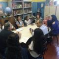 Representantes de secretarias, da escola e alunos estão sentados em torno de uma mesa. Em uma das paredes está encostada uma estante de livros. #Pracegover