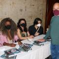 homem encosta em livros sobre a mesa observado por 3 mulheres #paratodosverem