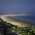 Imagem área da praia a noite, aparecendo prédios, jardins, faixa de areia e mar #paratodosverem