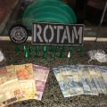 dinheiro e drogas sobre uma mesa com o distintivo da Rotam. #paratodosverem