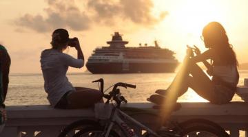 duas pessoas estão sentadas na mureta da orla assistindo à saída de um navio. Os dois fotografam com celulares. A foto foi feita ao por do sol, que cai e brilha do lado direito. #paratodosverem