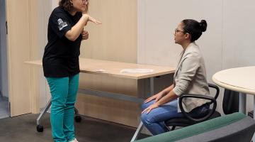Mulher em pé faz gesto em Libras para outra mulher que está sentada. #paratodosverem 