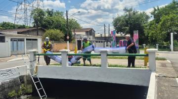 homens estão do lado de fora do canal segurando barreira e preparando para colocar na água. #paratodosverem 