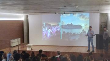 pessoas estão sentadas em auditório assistindo homem que fala diante de uma projeção. #paratodosverem