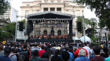 Público está na praça Mauá, diante do grande palco ao fundo e na frente da Prefeitura. A sinfônica toca. 