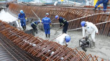 Sete homens trabalham com uniforme e capacete sobre estrutura de concreto e metal do viaduto em construção