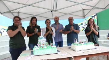 Quatro mulheres e dois homens batem palmas atrás de mesa com bolo de aniversário na cor verde