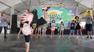 Em primeiro plano, menino faz acrobacia no chão. Atrás, onze crianças asssitem batendo palmas