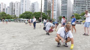 Pessoas caminham e se agacham na areia da praia. Elas fazem movimentos para coletar partículas pequenas de lixo. Ao fundo se vê os prédios da orla. #Pracegover