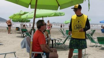 Agente uniformizado conversa com banhista que está sentado em cadeira na praia. Ao lado há apetrechos para o descarte correto de lixo. #Pracegover
