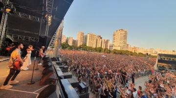 Músicos no palco e imensa multidão no chão, com vista a partir da praia para dentro da cidade. #Pracegover