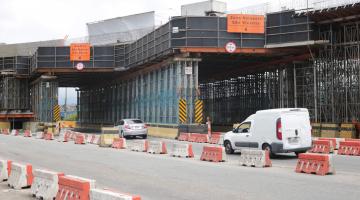 Grande estrutura em ferro que sustenta a obra de viaduto. Carros passam pista abaixo da obra. #Paratodosverem