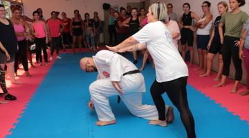 aula de defesa pessoal, mulher empurra homem pelas costas. Ele está com um dos joelhos no chão. Alunas em pé e em torno assistem à demonstração. #paratodosverem 