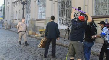 gravação de filme no centro histórico. Homem fardado caminha sobre paralelepípedo. Câmeras e microfones à frente. #paratodosverem 