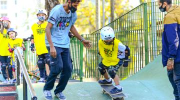 Criança andando de skate desce rampa sendo orientada por professor. #paratodosverem