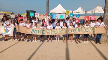alunos posam para foto na faixa de areia da praia segurando uma faixa onde se mudanças climáticas. #paratodosverem 