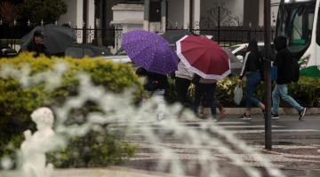 pessoas estão andando na rua de guarda chuva. Em primeiro plano se vê os jatos de água de uma fonte e um trecho de jardim. #paratodosverem