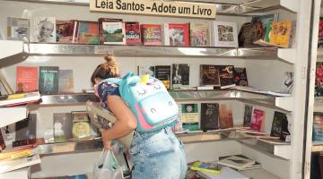 mulher com mochila nas costas está mexendo em livros em prateleira. #paratodosverem