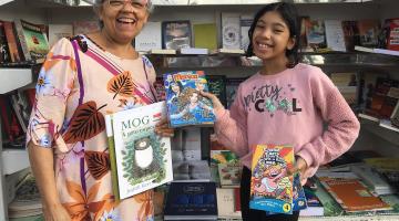 mulher e menina posam para foto segurando livros e gibis. Ao fundo, uma prateleira cheia de livros. #paratodosverem 