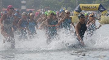 Mulheres entram no mar. A imagem mostra inúmeras mulheres e a água de mexendo com o movimento das atletas. #Paratodosverem