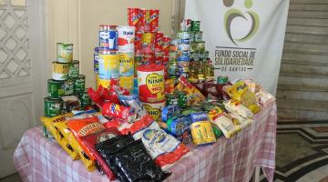 mesa com alimentos não perecíveis empilhados.Ao fundo, um banner do fundo social de solidariedade. #paratodosverem