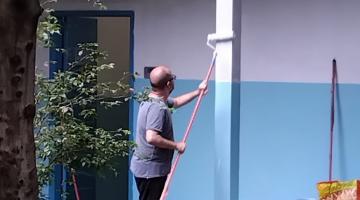 homem segura rodo de pintura com cabo longo e pinta coluna externa de escola. #paratodosverem 