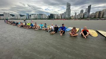 alunos estão sentados na areia em aquecimento para a aula. todos estão com pranchas. #paratodosverem