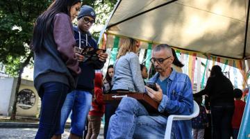 O cartunista desenha caricatura sentado em uma cadeira na praça. Há dois jovens na frente dele. #Pracegover