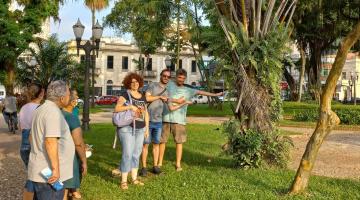 grupo de pessoas está em praça sobre a grama e com árvores em volta. #paratodosverem