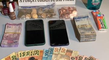 Mesa com cédulas de dinheiro, moedas, celulares e um shampoo. #paratodosverem