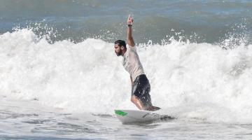surfista está sobre a prancha no mar. Seu braço esquerdo está para o alto. #paratodosverem 