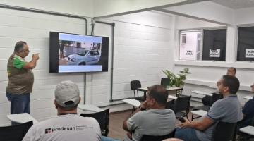 homem está em pé ao lado de um telão dando palestra para pessoas sentadas num pequeno auditório. #paratodosverem