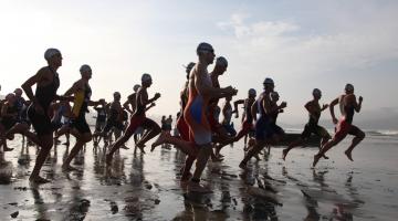 Atletas correm em direção a água na praia. #paratodosverem