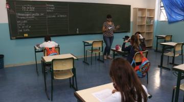 Em uma sala de aula, professora em frente a lousa e alunos nas carteiras escrevendo. #paratodosverem