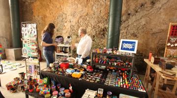 feira de artesanato com mesas cheias de pequenas peças. Duas pessoas estão em pé em torno dessas mesas. #paratodosverem 