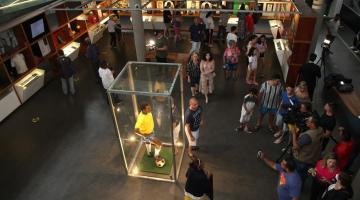 escultura de pelé dentro de redoma de vidro e vários visitantes circulando dentro de museu. #paratodosverem