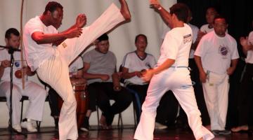 dois homens jogam capoeira. Músicos estão ao fundo. #paratodosverem