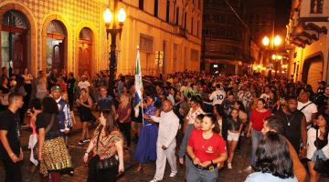 grande volume de pessoas caminha por rua do centro histórico à noite. Os postes de modelo republicano estão acesos. #paratodosverem 