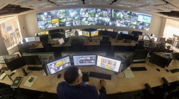 vista geral pelo alto do Centro de controle operacional, com um homem operando três computadores em primeiro plano e várias telas ao fundo. #paratodosverem