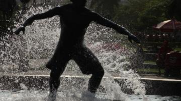 estátua do surfista contra a luz, parecendo uma sombra, com água jorrando sobre o atleta. #paratodosverem 