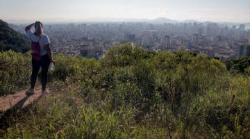 mulher está em alto de morro olhando para o horizonte. ao fundo se vê a cidade. #paratodosverem 